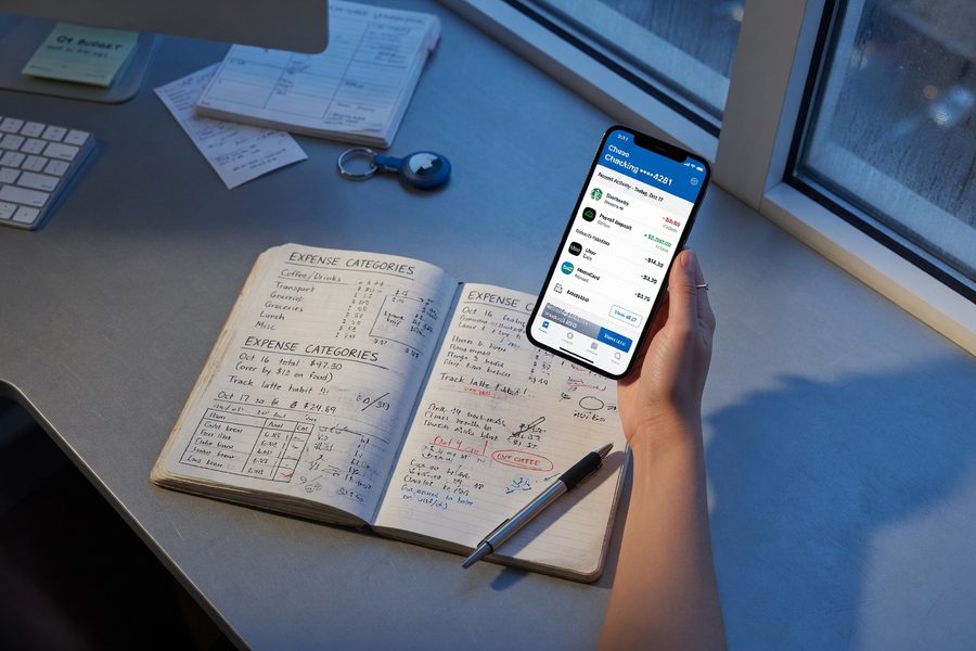 Person reviewing bank app transaction history on phone with a notebook open for tracking spending patterns